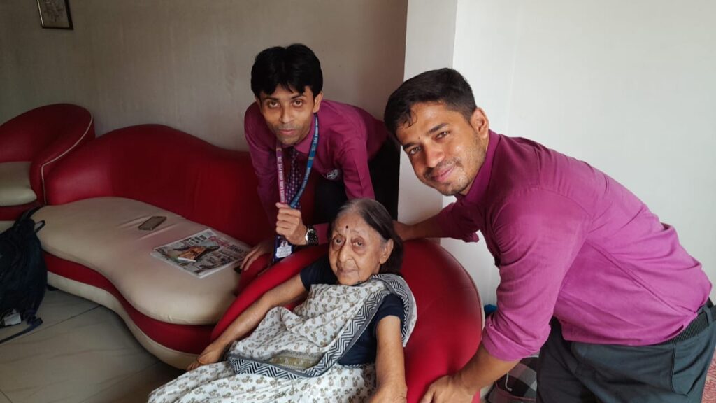 A smiling elderly woman sitting with a caregiver in a cozy Kolkata home; caregiver offering tea and gentle conversation